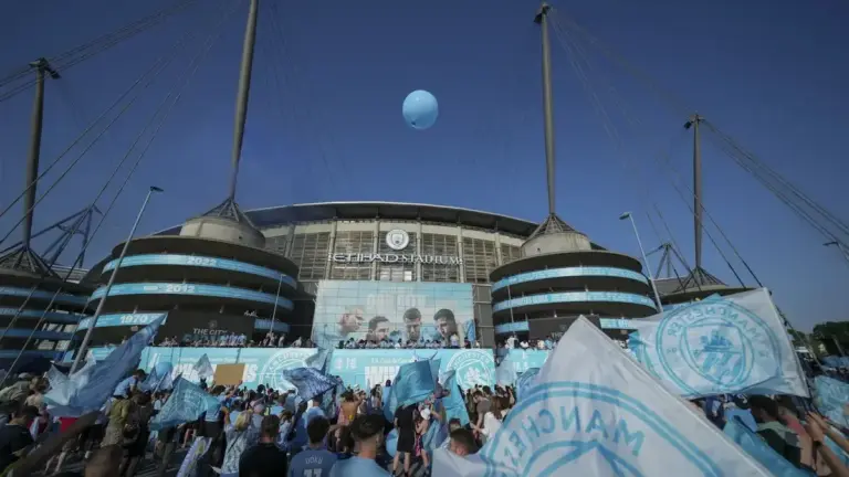 Man City 115 charges response: An exterior wide shot of the Etihad Stadium with Manchester City branding and supporters waving flags outside.